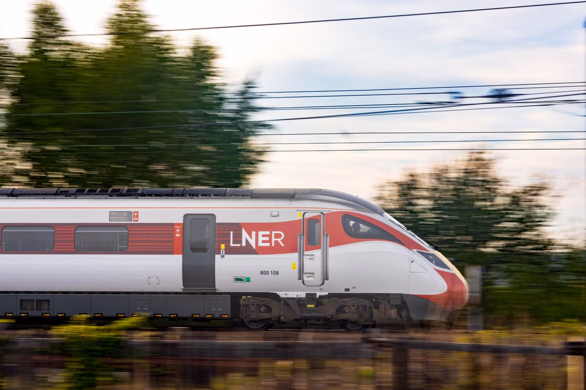 A red and white LNER Azuma train passing at speed under overhead wires, with blurred trees in the background.
