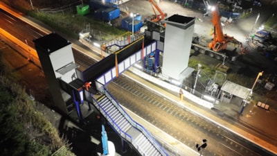 A steel footbridge span being lifted by crane over railway tracks at a station
