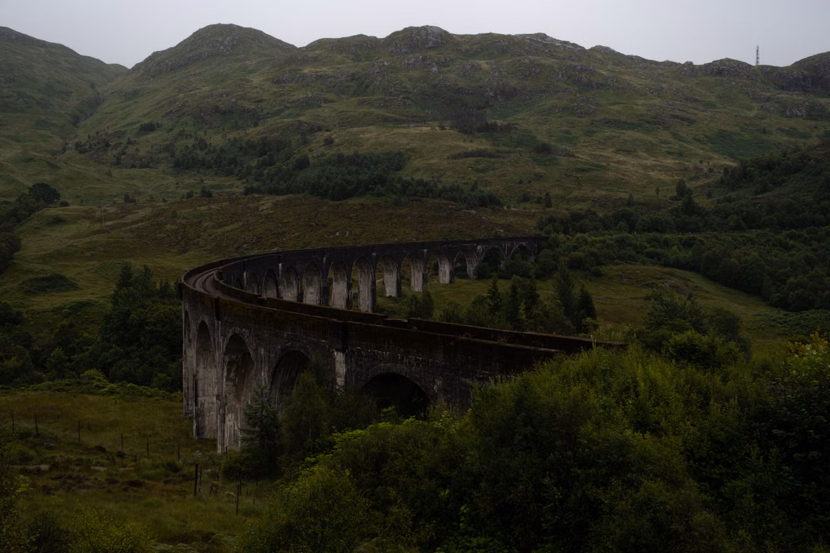 A curved stone railway viaduct with many arches crossing a green valley below grassy hills on an overcast day.