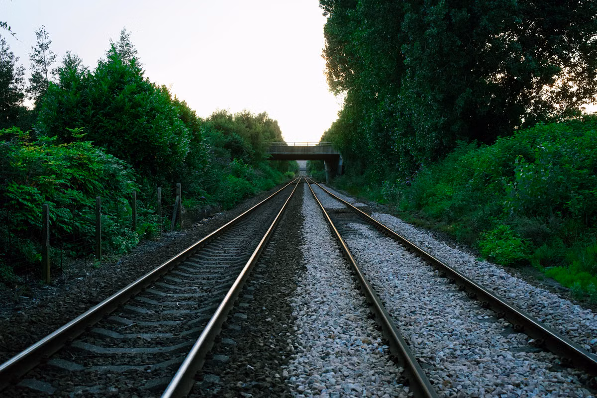 A straight stretch of double-track railway on ballast running between green trees and bushes towards a road bridge in the distance.