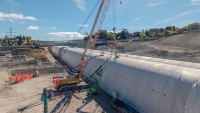 A crane lifting a precast concrete arch segment into place on a cut-and-cover tunnel under construction